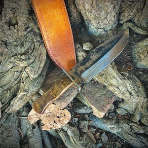 May include: A handcrafted hunting knife with a wooden handle and brass rivets, resting on a bed of textured tree roots and rocks. The knife has a dark, weathered blade and is accompanied by a tan leather sheath.