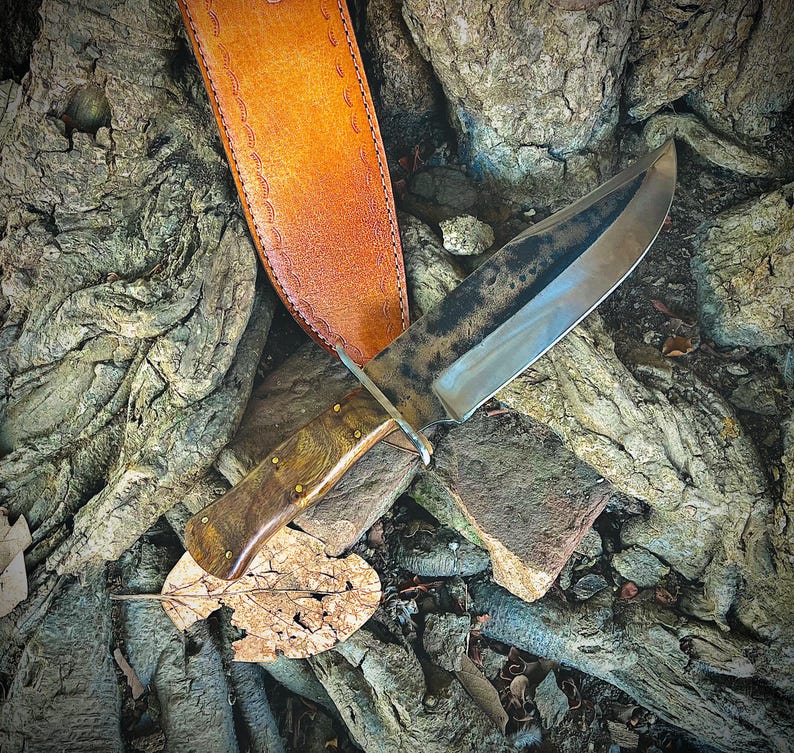 May include: A handcrafted hunting knife with a wooden handle and brass rivets, resting on a bed of textured tree roots and rocks. The knife has a dark, weathered blade and is accompanied by a tan leather sheath.