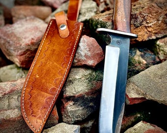 May include: A large, polished steel knife with a dark wooden handle and a leather sheath. The sheath is brown with decorative stitching and a strap. The knife is resting on a pile of red and brown rocks.