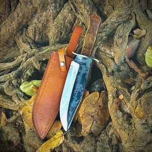 May include: A large, handcrafted hunting knife with a wooden handle and a leather sheath. The blade has a dark, textured finish. The sheath is brown and has a strap with a button closure. The knife and sheath are resting on a bed of leaves and tree roots.