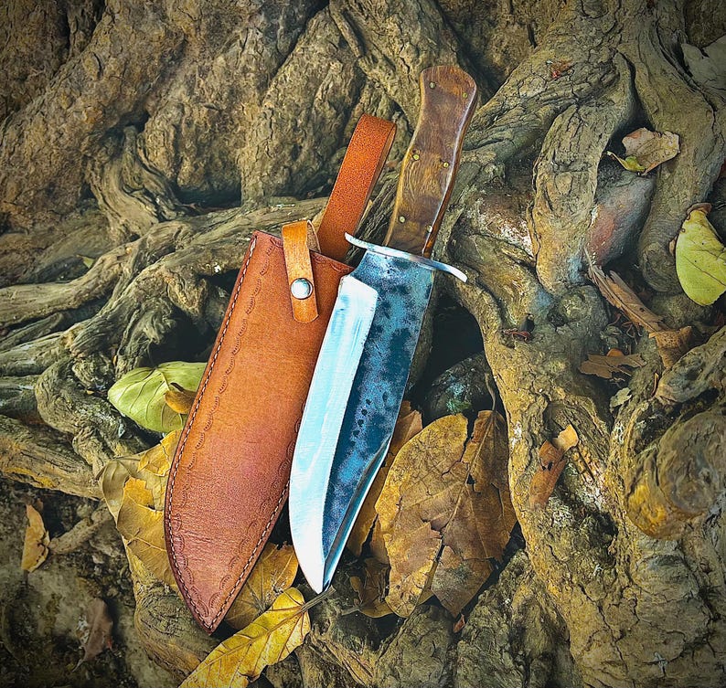 May include: A large, handcrafted hunting knife with a wooden handle and a leather sheath. The blade has a dark, textured finish. The sheath is brown and has a strap with a button closure. The knife and sheath are resting on a bed of leaves and tree roots.