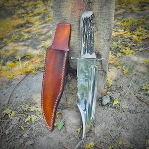 May include: A large hunting knife with a polished, serrated blade and a handle made of deer antler. The knife is next to a brown leather sheath with white stitching. The knife and sheath are leaning against a tree trunk.