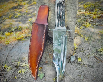 May include: A large hunting knife with a polished, serrated blade and a handle made of deer antler. The knife is next to a brown leather sheath with white stitching. The knife and sheath are leaning against a tree trunk.
