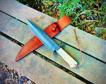 May include: A large, silver-bladed knife with a cream-colored handle and a brown leather sheath. The knife has a decorative design on the blade and is resting on a wooden surface.