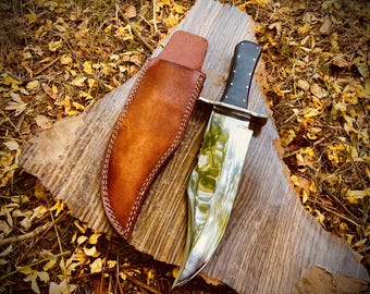 May include: A large, polished steel knife with a black handle and a brown leather sheath. The knife blade reflects the surrounding environment. The sheath is stitched with white thread. The knife and sheath rest on a weathered wooden surface.