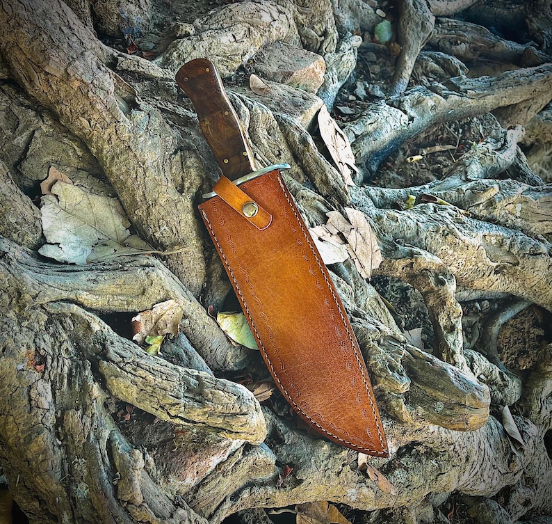 May include: A large hunting knife with a wooden handle and a leather sheath. The knife has a silver blade and is secured in a brown leather sheath with a button closure. The knife and sheath are resting on a bed of tree roots.
