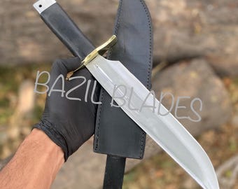 May include: A large, polished steel knife with a black handle and a brass bolster, accompanied by a black leather sheath. The knife has a straight blade and the text "BAZIL BLADES" is visible.