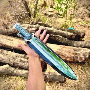 May include: A dagger with a polished, reflective blade and a blue-grey handle. The dagger is in a black leather sheath with white stitching. The handle has a series of holes. The dagger is held in front of a background of logs and foliage.