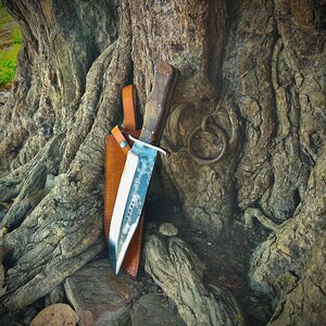 May include: A large, handmade knife with a wooden handle and a polished steel blade. The knife is sheathed in a brown leather case and rests against a textured tree trunk. The blade has a unique, worn appearance. A metal ring is attached to the tree.