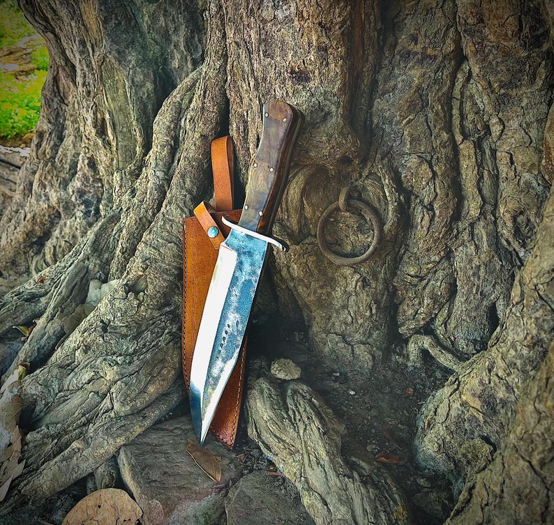 May include: A large, handmade knife with a wooden handle and a polished steel blade. The knife is sheathed in a brown leather case and rests against a textured tree trunk. The blade has a unique, worn appearance. A metal ring is attached to the tree.
