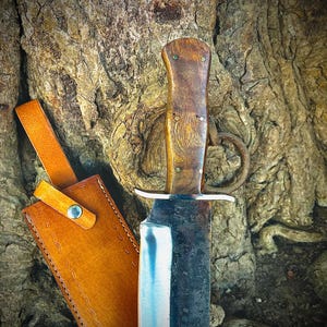May include: A handcrafted knife with a wooden handle and a leather sheath. The blade is partially visible, with a silver edge and a dark, textured surface. The sheath is tan with a strap and a button closure. The knife is displayed against a tree trunk.
