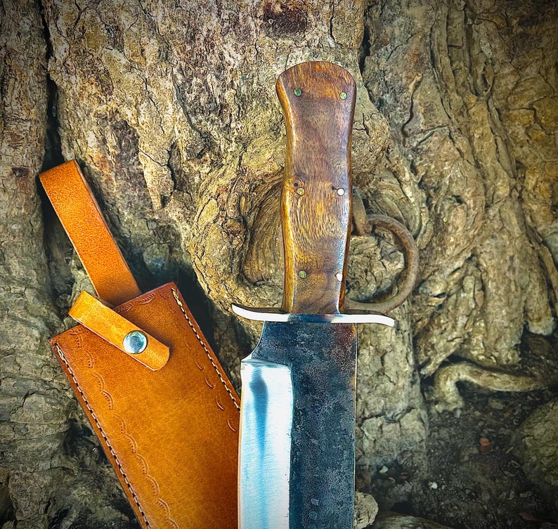 May include: A handcrafted knife with a wooden handle and a leather sheath. The blade is partially visible, with a silver edge and a dark, textured surface. The sheath is tan with a strap and a button closure. The knife is displayed against a tree trunk.
