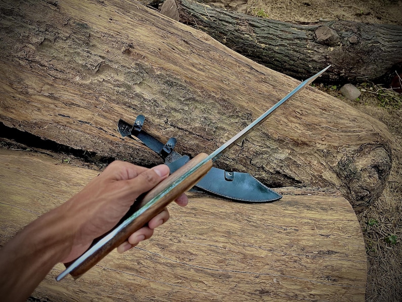A handmade machete knife displayed with its leather sheath. Crafted from 1095 steel with a full tang for strength, this large knife is suitable for survival and camping. Makes a thoughtful and rugged gift for husbands, fathers, or any outdoorsman who appreciates handmade craftsmanship.