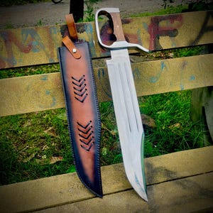 Può includere: Un grande coltello con lama argentata, manico in legno e fodero in pelle. Il coltello ha un'impugnatura curva distintiva e una lama larga. Il fodero è marrone e nero con cuciture decorative. Il coltello è progettato per uso esterno.
