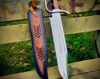 May include: A large, silver-bladed knife with a wooden handle and a leather sheath. The knife has a distinctive curved handle and a wide blade. The sheath is brown and black with decorative stitching. The knife is designed for outdoor use.