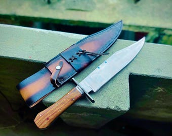May include: A large, handmade hunting knife with a polished steel blade and a wooden handle. The knife is accompanied by a brown leather sheath with decorative stitching and a leather strap. The sheath has a gradient of brown and black colors.