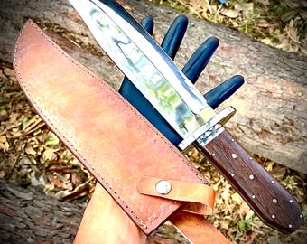 May include: A large, polished steel knife with a wooden handle and a leather sheath. The knife has a wide blade and a silver bolster. The handle is dark brown with decorative silver studs. The sheath is tan leather with a strap and snap closure.