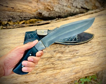 May include: A large, silver-colored knife with a black handle and a leather sheath. The knife is held in a hand, with the blade facing upwards. The sheath is black with a decorative pattern.