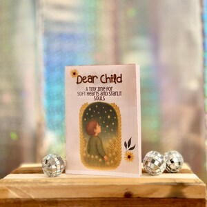 May include: A zine titled "Dear Child" with a drawing of a child looking up at stars. The cover has text that reads "A tiny zine for soft hearts and starlit souls." Two disco balls sit on a wooden surface.