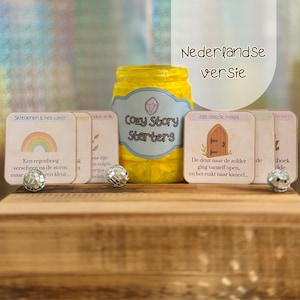 May include: A yellow jar labeled "Cozy Story Starters" with a light blue label and a crystal graphic. Several small cards with text and illustrations are in front of the jar. Two decorative silver balls are on the wooden surface.