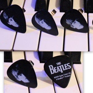 Puede incluir: Cuatro púas de guitarra negras con imágenes de los miembros de la banda The Beatles. Las púas tienen un anillo plateado en la parte superior. Una púa tiene el texto "The Beatles" y la información de copyright "©2009 Apple Corps Ltd. A Beatles™ Product".