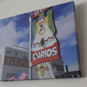 May include: A vintage neon sign with a teepee design and the words "Curios" in red letters. The sign is mounted on a pole and has a blue sky in the background. The sign also features a Native American symbol.