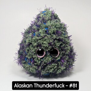 May include: A plush, rounded creature with large, black eyes and a textured, fuzzy body. The body is primarily green with blue and purple accents. The text "Alaskan Thunderfuck - #81" is displayed in a black rectangle at the bottom.
