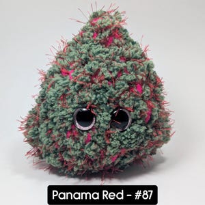 May include: A handmade plush toy in a teardrop shape, featuring a textured green and pink yarn. The toy has large, round, black eyes and red accents. The text "Panama Red - #87" is displayed on a black rectangle.