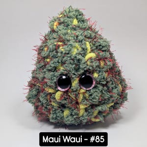 May include: A handmade, plush creature with large, round, black eyes and a fuzzy, textured body. The body is primarily green with yellow and red accents. The text "Maui Waui - #85" is displayed on a black rectangle at the bottom.