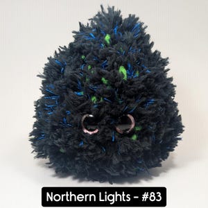 May include: A plush, round creature with black, blue, and green textured fur. It has large, round eyes with pink accents. The text "Northern Lights - #83" is displayed in a black rectangle at the bottom.