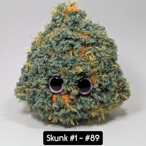 May include: A handmade plush skunk toy with large, round eyes. The skunk is made of green, orange, and gold yarn. The text "Skunk #1 - #89" is displayed on a black rectangle at the bottom of the image.