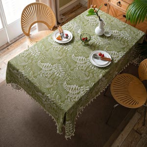 Può includere: Una tovaglia verde con un motivo a foglie bianche copre un tavolo di legno. La tovaglia ha un bordo con frange. Due sedie di legno sono posizionate vicino al tavolo. Piatti, posate e un vaso con fiori sono sul tavolo.