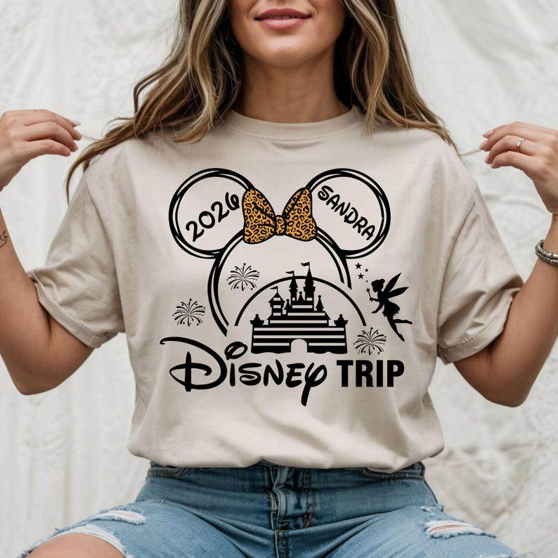 May include: Beige t-shirt with a black graphic featuring Mickey Mouse ears, a leopard print bow, a castle, and the words "Disney Trip". The ears have the text "2026" and "Sandra". A fairy and fireworks are also included.