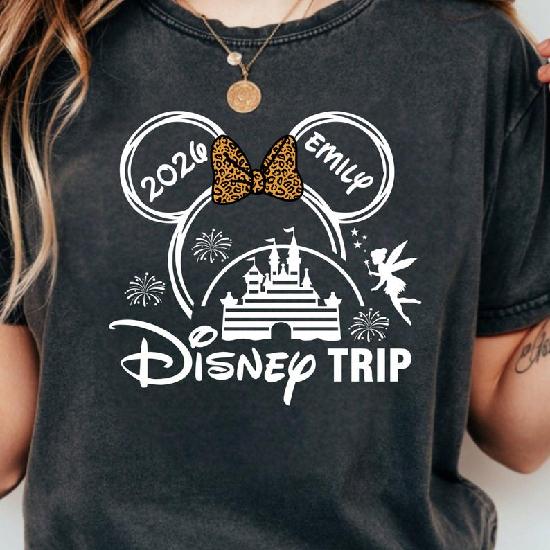 May include: Dark gray t-shirt with a white Disney Trip design. The design includes Mickey Mouse ears, a leopard print bow, the name "Emily", the year "2026", a castle, a fairy, and fireworks.