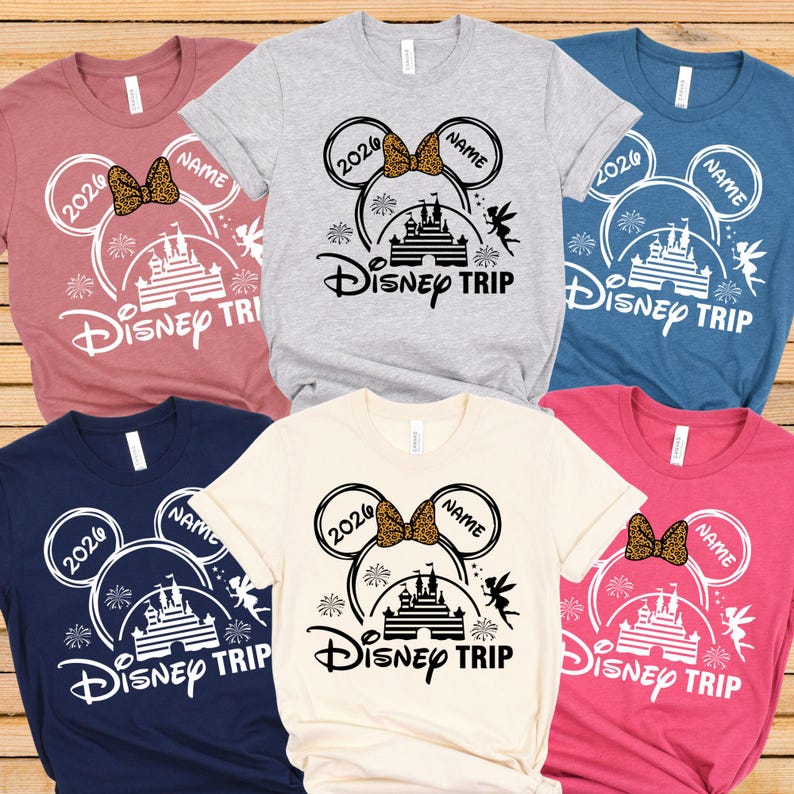 May include: Assorted-color t-shirts with a Disney Trip design. Each shirt features a Mickey Mouse ear design with the year 2026, a bow, and the word "NAME". The design includes a castle, fireworks, and the words "Disney Trip".