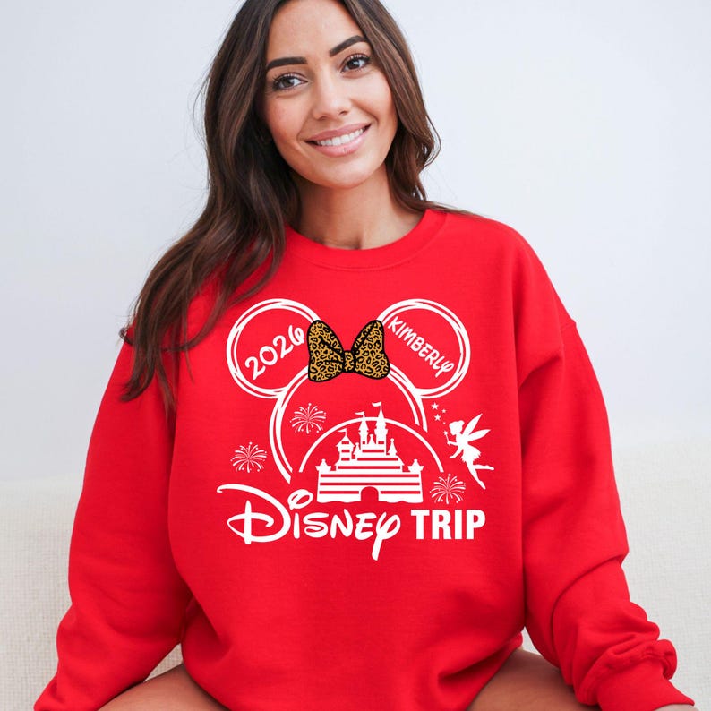 May include: Red sweatshirt with a white Disney Trip graphic. The design includes Mickey Mouse ears with the year 2026 and the name "Kimberly", a castle, fireworks, and a fairy. A leopard print bow is in the center.