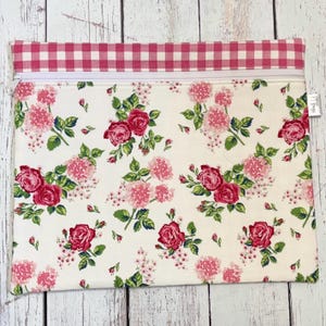 May include: A rectangular zippered pouch with a floral pattern of pink roses and hydrangeas on a cream background. The top border is a pink and white gingham check. The pouch has a white zipper.