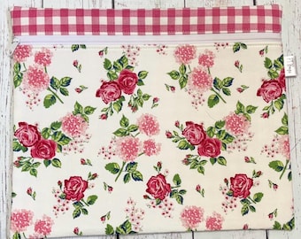 May include: A rectangular zippered pouch with a floral pattern of pink roses and hydrangeas on a cream background. The top border is a pink and white gingham check. The pouch has a white zipper.