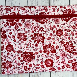 May include: A white zippered pouch with a red floral pattern. The pattern includes various flowers, leaves, and butterflies. The zipper is a deep red color, and the pouch is rectangular.