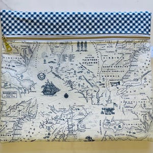 May include: A rectangular pouch featuring a map of the Thirteen Colonies in blue and white. The pouch has a blue and white gingham pattern at the top, a gold zipper, and a small tag. The map includes illustrations of ships and historical text.