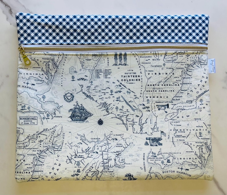 May include: A rectangular pouch featuring a map of the Thirteen Colonies in blue and white. The pouch has a blue and white gingham pattern at the top, a gold zipper, and a small tag. The map includes illustrations of ships and historical text.