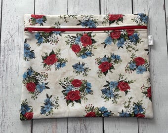 May include: A rectangular cream-colored zippered pouch with a floral pattern. The pattern features clusters of red roses and blue flowers with green foliage. The zipper is red.