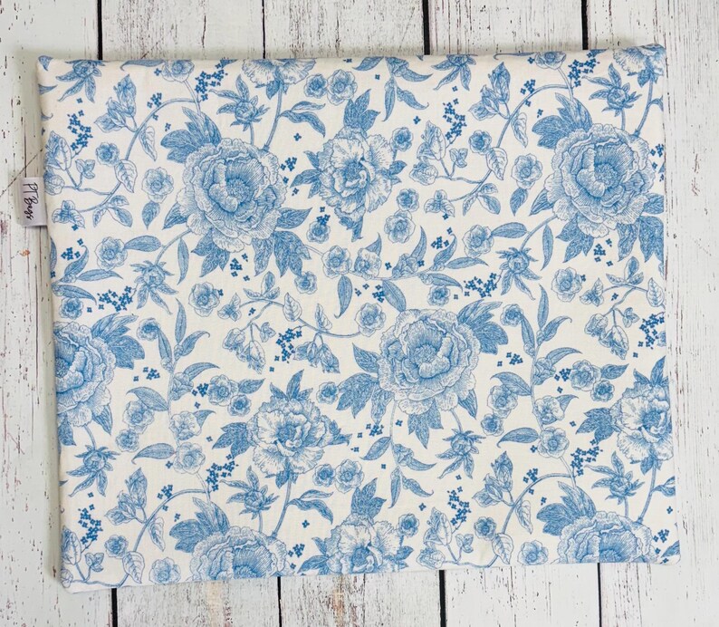 May include: A rectangular fabric item with a floral pattern. The design features large, light blue peony flowers and smaller floral accents on a white background. The overall aesthetic is reminiscent of a vintage toile design.
