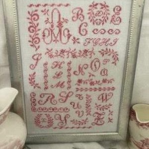 May include: Framed cross-stitch sampler in a silver-colored frame. The sampler features red embroidered letters and floral designs on a white background. A decorative piece with a vintage aesthetic.