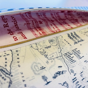 May include: Open pencil case with a gold zipper. The interior lining features red musical notes and the text "to the Republic and justice." The exterior displays a map of the Thirteen Colonies.