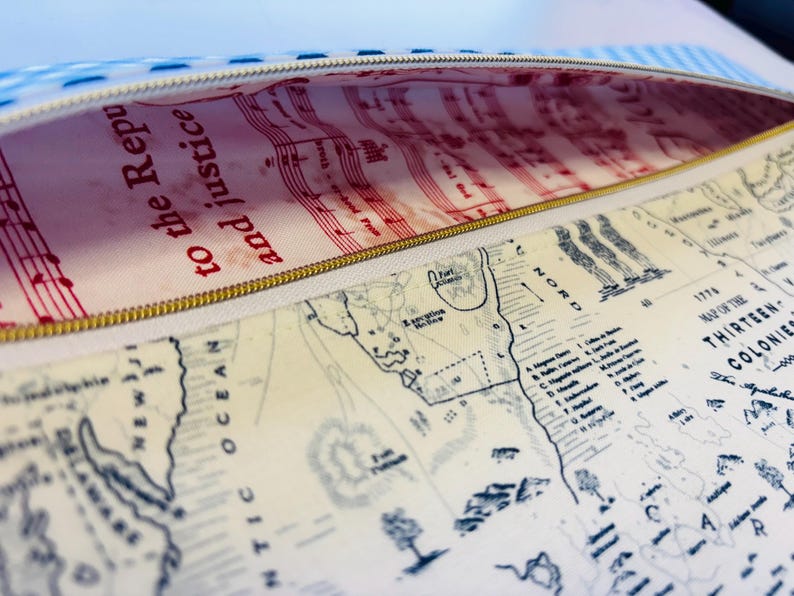 May include: Open pencil case with a gold zipper. The interior lining features red musical notes and the text "to the Republic and justice." The exterior displays a map of the Thirteen Colonies.