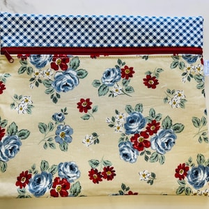 May include: A rectangular fabric pouch with a floral pattern. The pouch features a cream-colored background with blue and red roses. The top section is blue and white checkered fabric, separated by a red zipper.