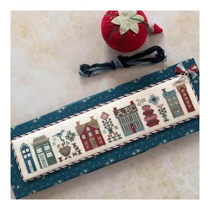May include: A cross-stitch sampler featuring a row of colorful houses with intricate details. The sampler is on a blue fabric base, with a red strawberry pin cushion and black embroidery floss nearby. The houses are in shades of red, blue, and green.