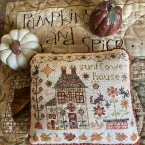 May include: A cross-stitch embroidery of a brown house with a sunflower design and the text "sunflower house". The house has a red door and white windows. The embroidery is surrounded by a brown border.