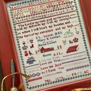 May include: A framed cross-stitch sampler with a red frame. The sampler features embroidered text and images in various colors, including red, green, and blue. The text includes phrases like "The Lord is my rock" and "Love me Ever." A bell and measuring tape are in the foreground.